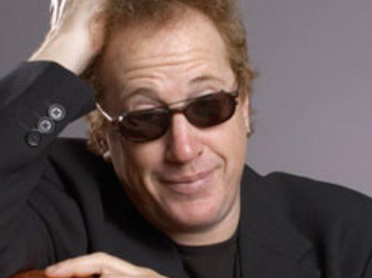 A man wearing sunglasses and exhibiting superior vocal health while holding an orange guitar.